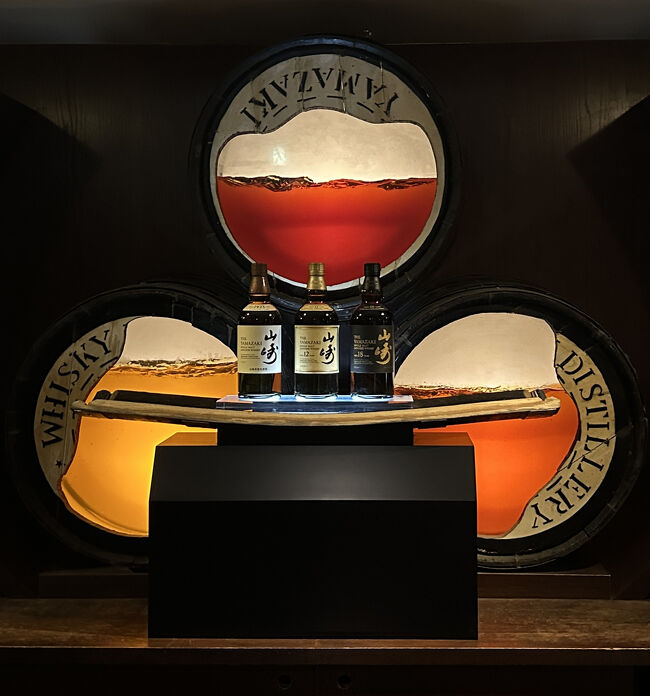 日比谷BAR WHISKY-S・バーカウンター<br />「ウイスキー天使の分け前」とは、ウイスキーの熟成中に、樽の中から自然と蒸発していくお酒のことを指します。まるで、樽の中に住む天使がそのお酒を少しずつ飲んでいるかのように見えることからこの名がつきました。ロマンチックで素敵な表現です。木製の樽は、私たちの目には密閉されているように見えても、実はミクロな穴がたくさんあります。樽の中のお酒は、その穴を通して少しずつ空気中に蒸発していきます。それを壁のオブジェで表現しています。<br />手前の架台には、山崎１８年・１２年・NVが置かれています。カッコイイです。